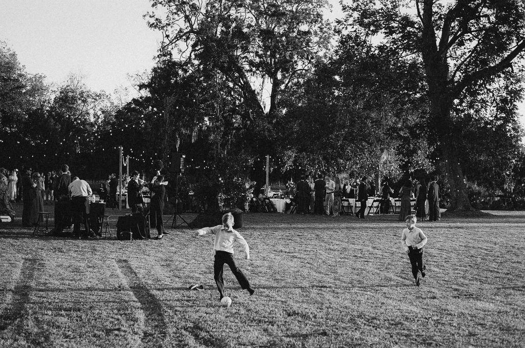 Black and white candid of kids playing soccer on the lawn during a wedding reception under the oaks