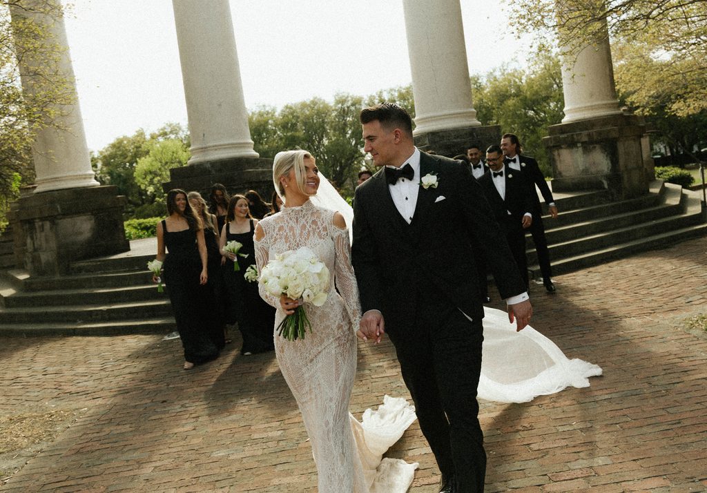 Bride and groom walking hand in hand with their wedding party past white columns on a spring day in Charleston