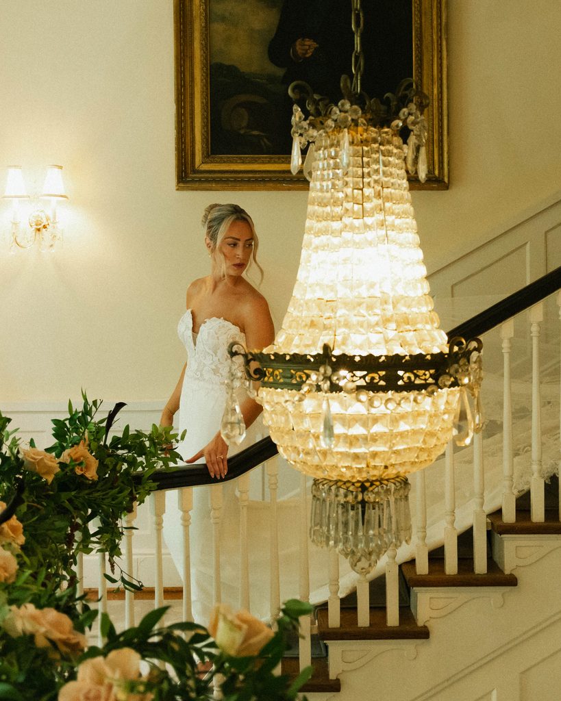 Bethany in her wedding gown on the grand staircase beside a crystal chandelier at The Morehead Inn in Charlotte, NC