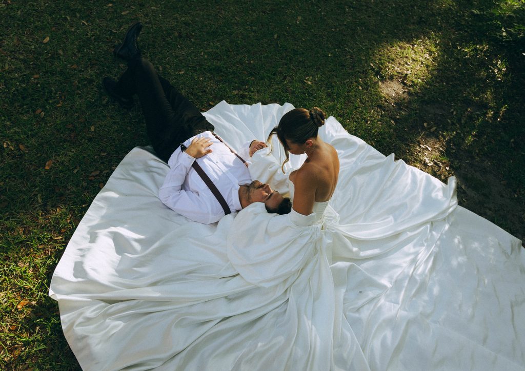 Bride and groom lying on the grass with her satin gown spread around them