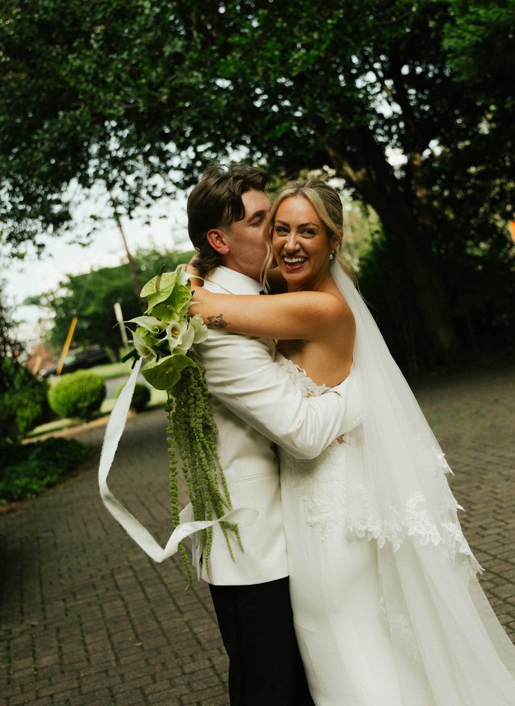 Ethan kissing Bethany as she laughs, holding a calla lily bouquet, in the garden at The Morehead Inn in Charlotte, NC