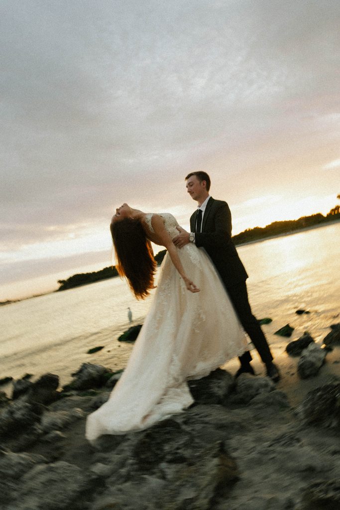 Groom dipping his bride on the rocks by the water at sunset