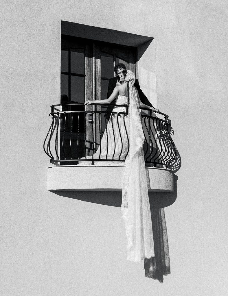 Black and white photo of a bride standing on a wrought iron balcony with her long veil hanging over the railing