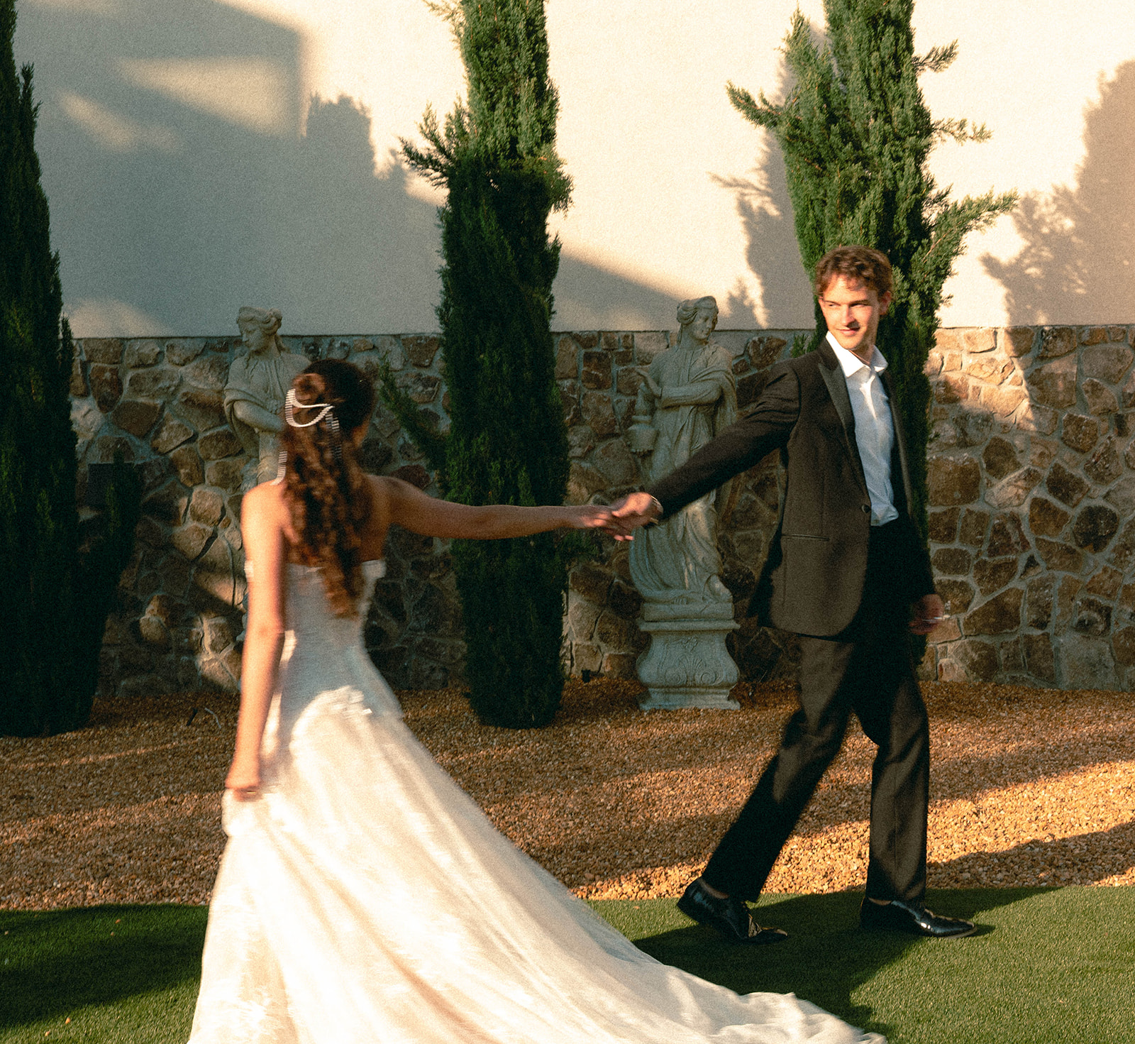 Bride leading the groom by the hand across a garden lawn past cypress trees and a stone wall in golden light