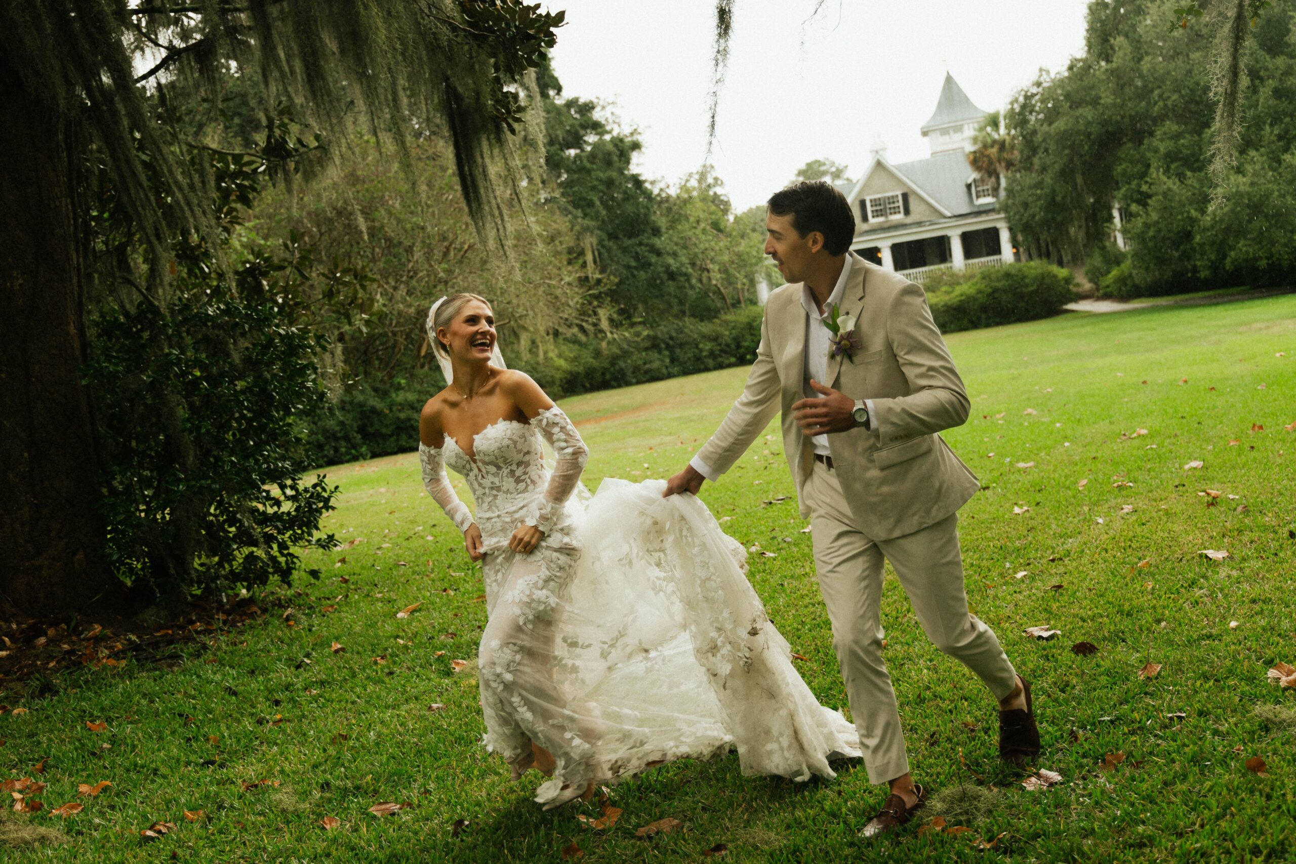Bride and groom laughing as they walk across a green lawn under oak trees with a historic white house behind them