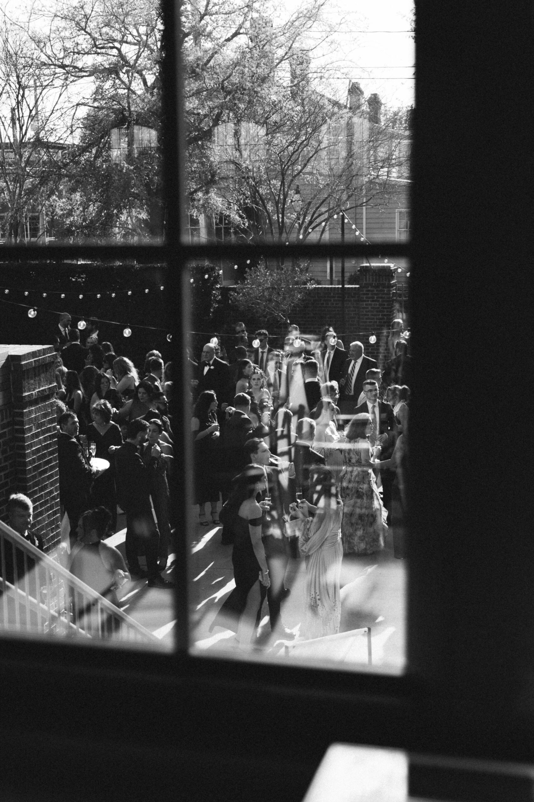 Black and white view through a window of guests celebrating at an outdoor wedding reception