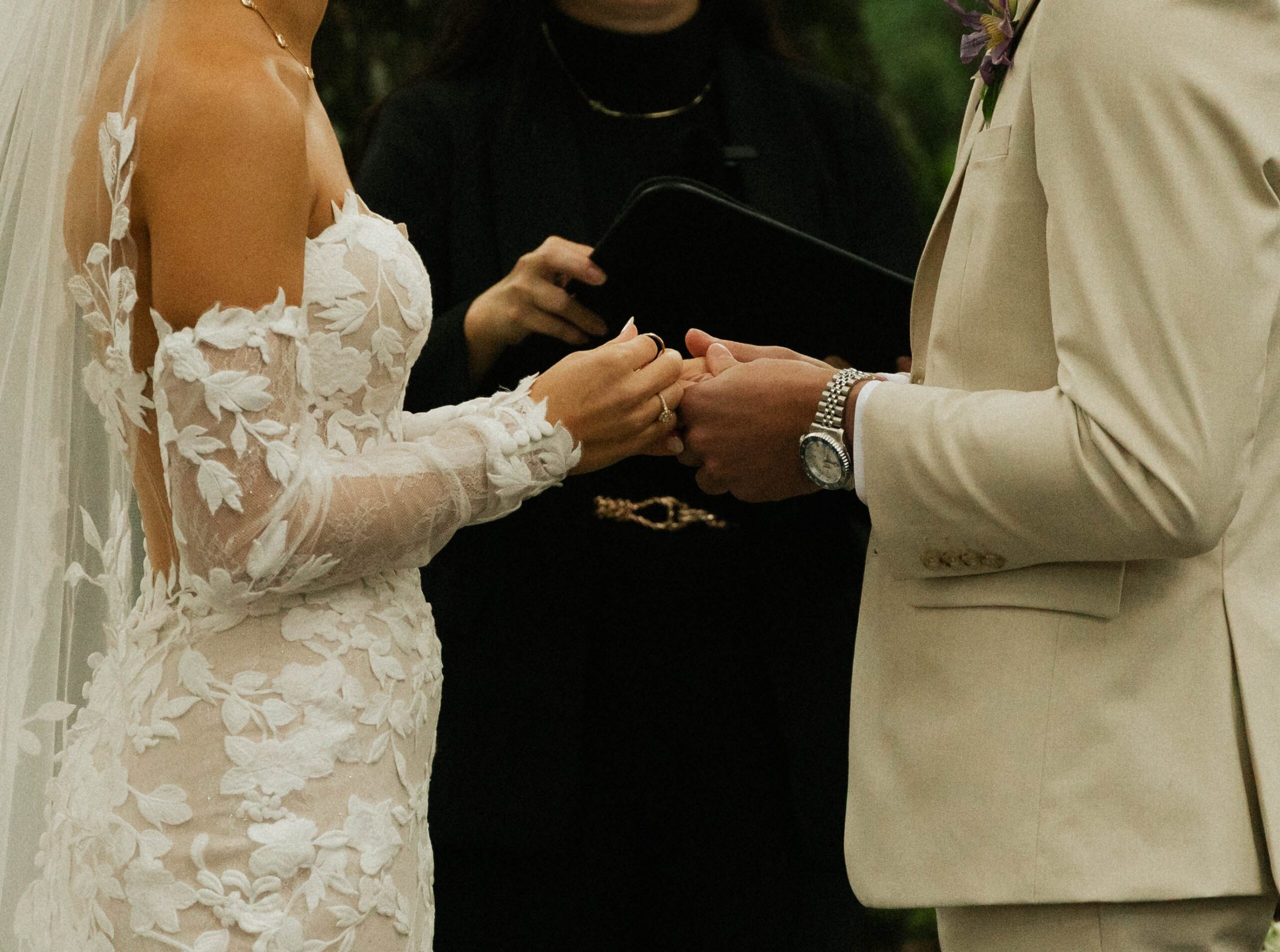 Bride in a lace off-the-shoulder gown and groom in a cream suit holding hands during their wedding ceremony