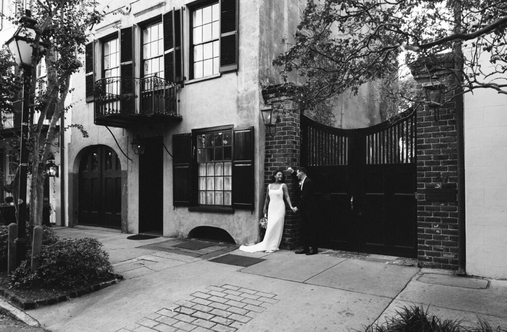 Black and white portrait of a bride and groom on a historic street beside a wrought iron gate, by a Charleston wedding photographer