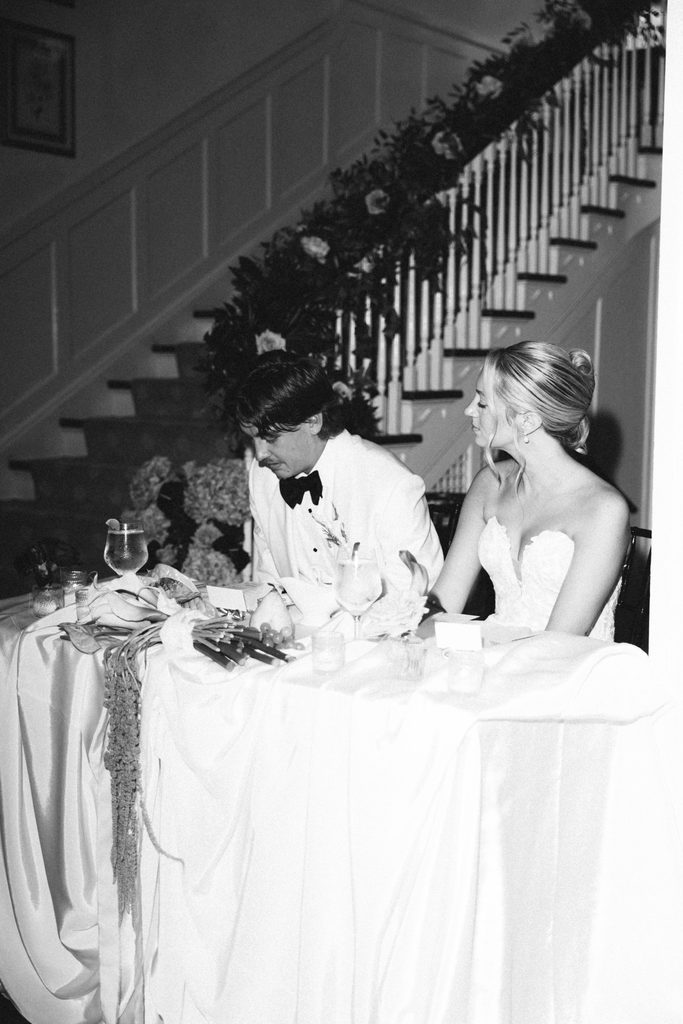 Black and white photo of Bethany and Ethan at their sweetheart table by the staircase at The Morehead Inn