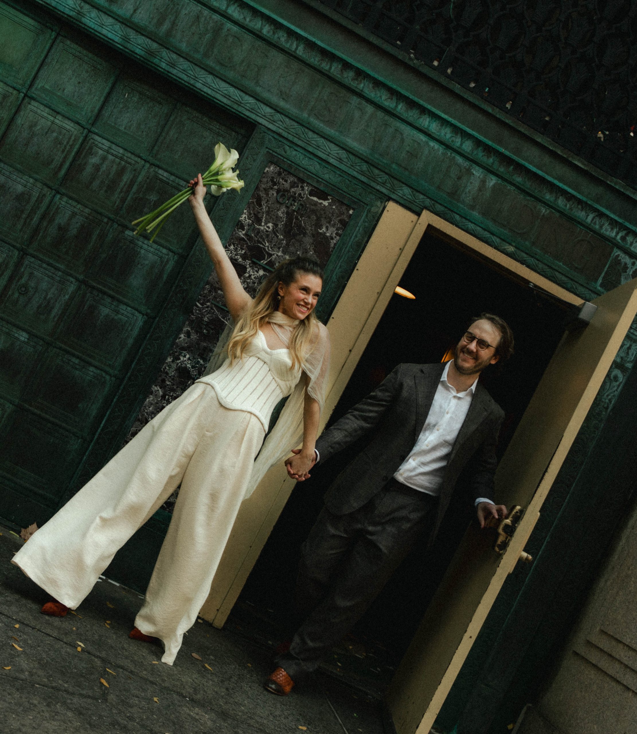 Lauren raising her calla lily bouquet in celebration as she and Jack walk out of a green doorway after their NYC elopement