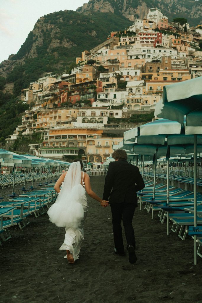 Bride and groom walking past beach umbrellas in Positano with the colorful cliffside village behind them