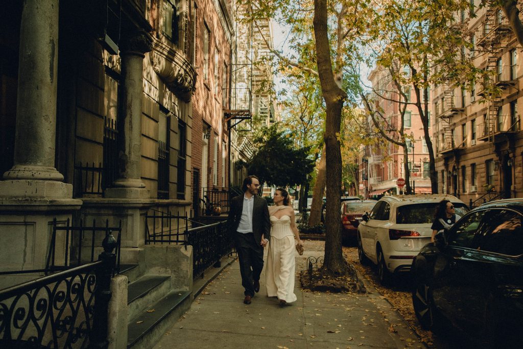 Lauren and Jack walking hand in hand down a tree-lined New York City sidewalk past brownstones
