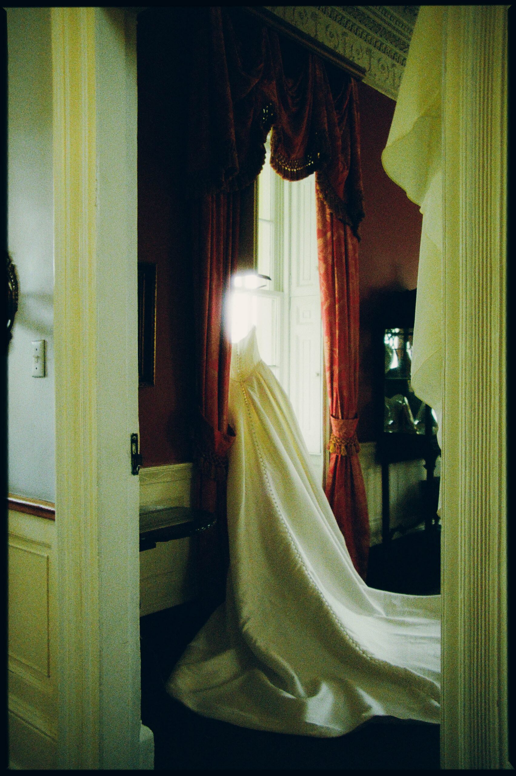 Bride in her wedding gown standing in a doorway with red curtains and soft window light, shot on 35mm film
