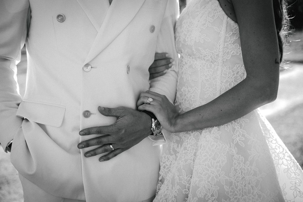Black and white close-up of a bride and groom holding each other, her hand with engagement ring resting on his cream suit