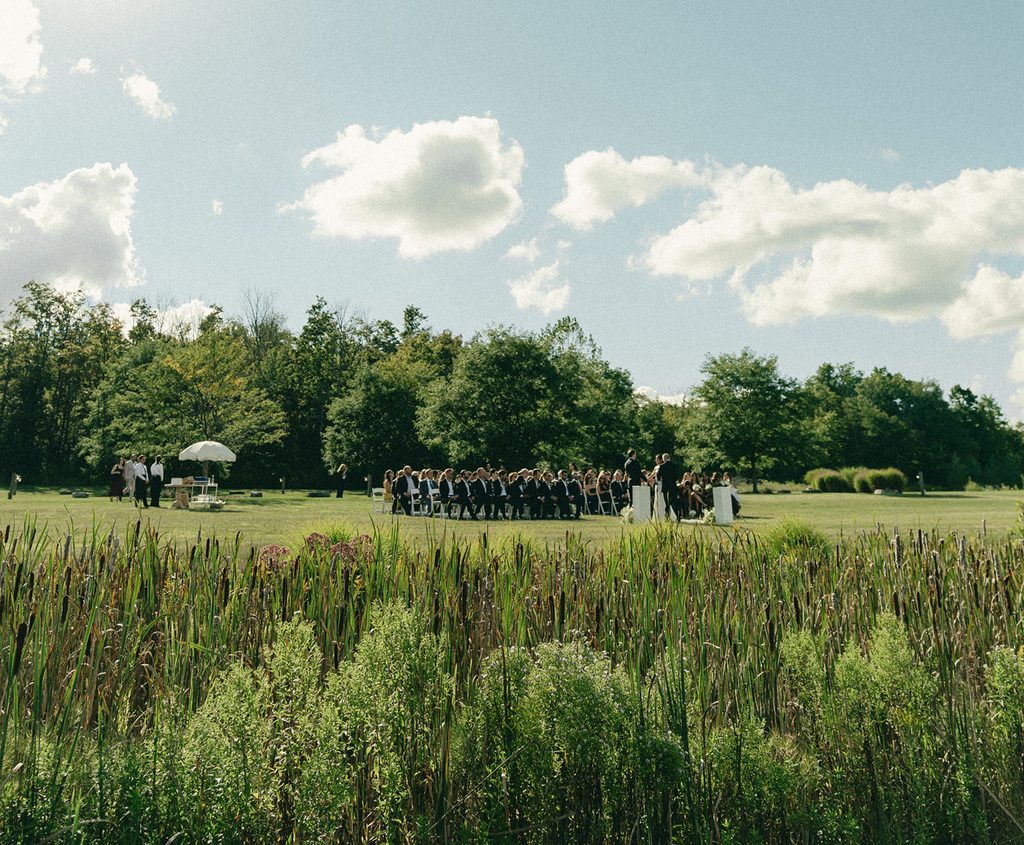 Wide view of an outdoor wedding ceremony on a green lawn surrounded by tall grasses and trees under a blue sky