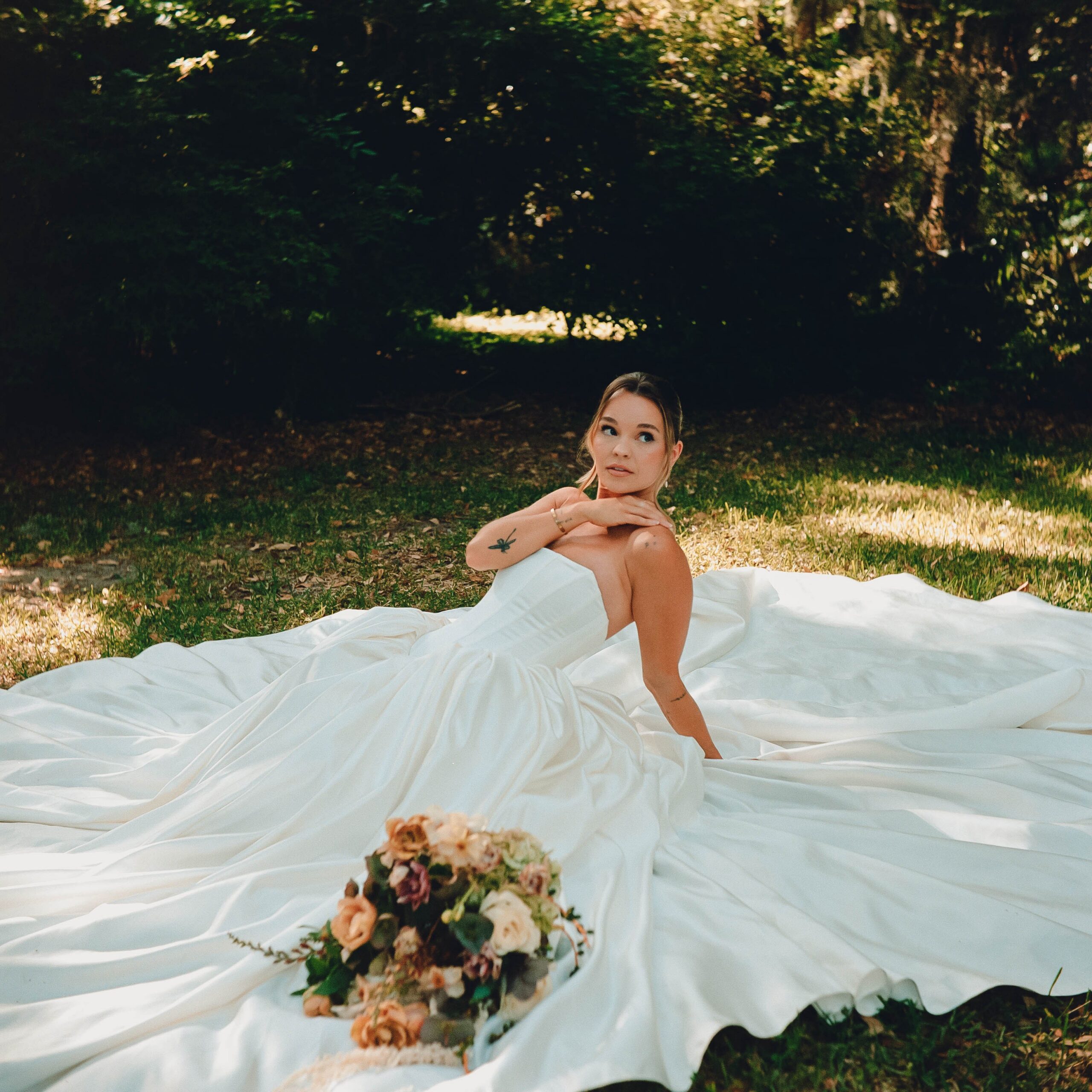Bride sitting in her flowing satin gown on the grass with her bouquet, a film wedding photography portrait
