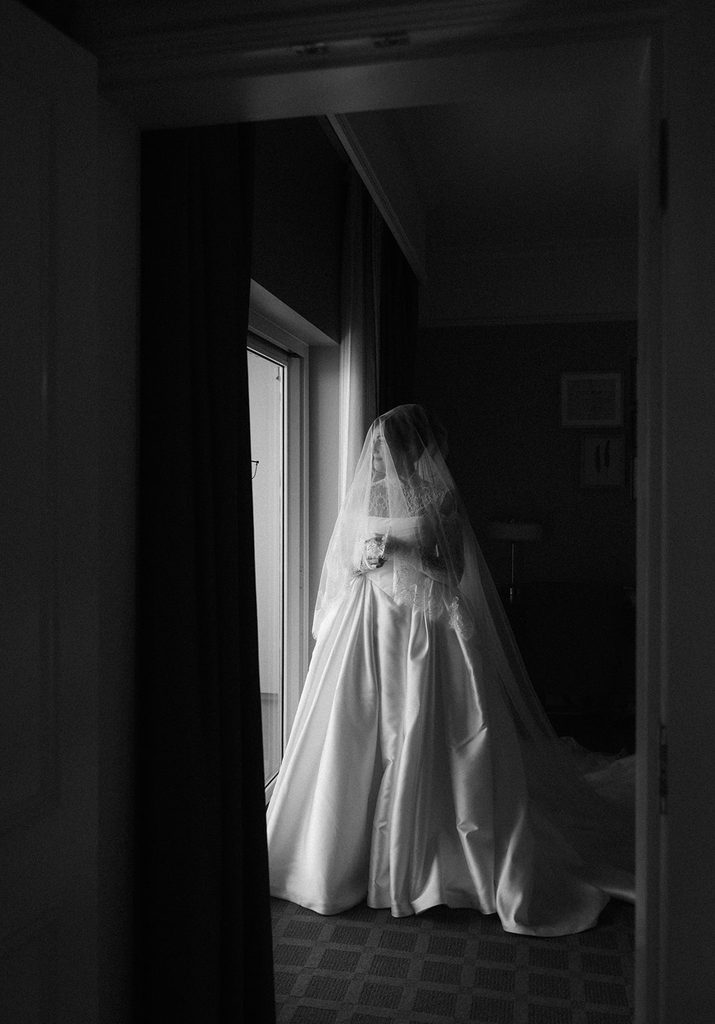 Black and white photo of a bride in her gown standing by a window while getting ready at The Ballantyne Hotel in Charlotte