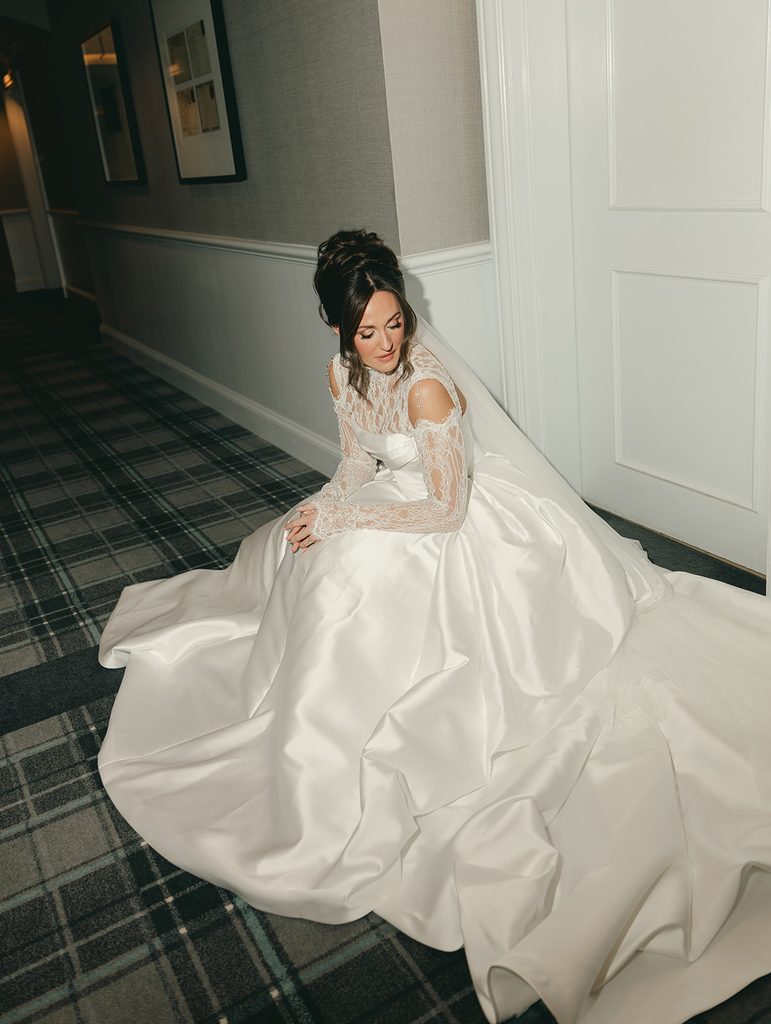 Bride in a lace wedding gown getting ready by a window on her wedding day